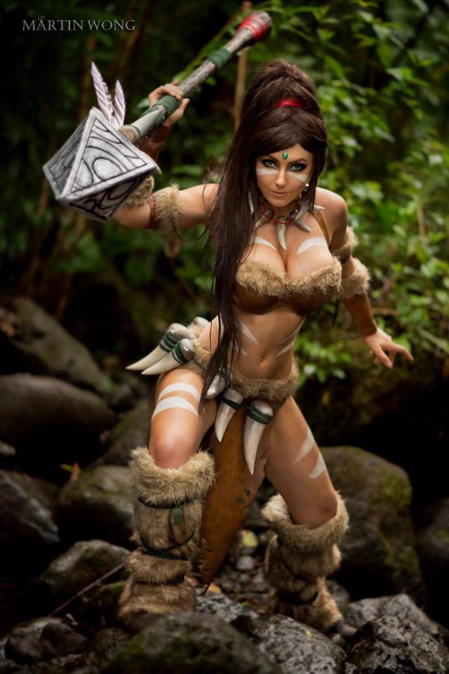 1girl boots bra breasts brown_hair chains cleavage collar cosplay fang jessica_nigri jungle league_of_legends long_hair looking_at_viewer nature nidalee outdoors photo ponytail skirt spear stone tan_skin tattoo underwear weapon