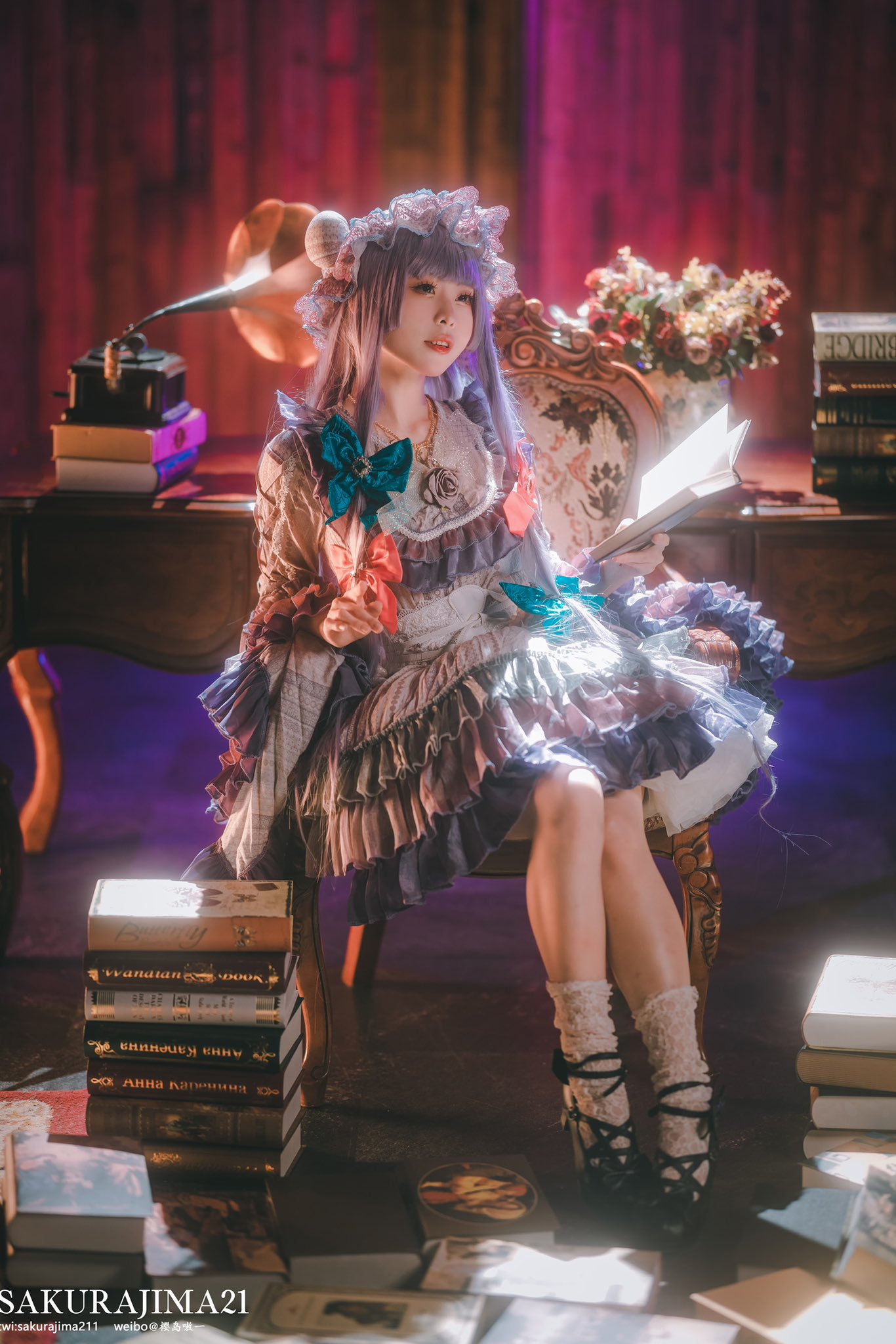 1girl aqua_bow artist_name black_footwear blunt_bangs blurry blurry_background book book_stack bouquet bow chair collarbone commentary cosplay_photo depth_of_field dress frilled_dress frills full_body hair_bow highres holding holding_book indoors long_sleeves on_chair open_book open_mouth patchouli_knowledge photo_(medium) purple_hair red_bow sakurajima211 socks solo symbol-only_commentary teeth touhou upper_teeth_only white_socks wooden_chair wooden_desk