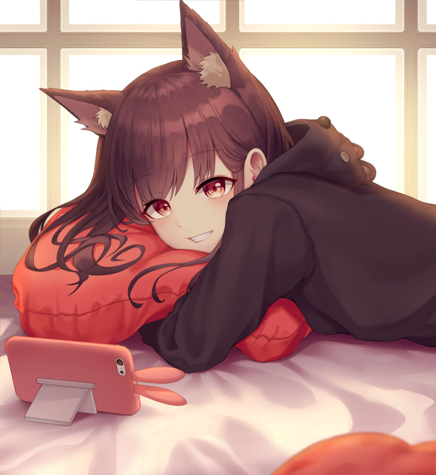 1girl :d animal_ear_fluff animal_ears bangs bear_hood bed_sheet black_hoodie blush brown_hair cat_ears cellphone eyebrows_visible_through_hair grin highres hood hood_down hoodie indoors long_hair long_sleeves lying myung on_bed on_stomach open_mouth original phone pillow pillow_hug red_eyes smartphone smartphone_case smile solo symbol_commentary teeth window