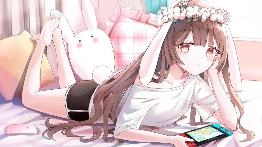 1girl :o animal_ears bare_shoulders barefoot bed_sheet black_shorts blush brown_eyes brown_hair bunny_girl bunny_tail cellphone collarbone commentary day flower flower_wreath frilled_pillow frills full_body head_wreath highres holding indoors iren_lovel legs_up long_hair looking_at_viewer lying nintendo_switch off_shoulder on_stomach original parted_lips phone pillow plaid rabbit_ears shirt short_shorts short_sleeves shorts soles solo stuffed_animal stuffed_bunny stuffed_toy sunlight symbol_commentary tail very_long_hair white_flower white_shirt window