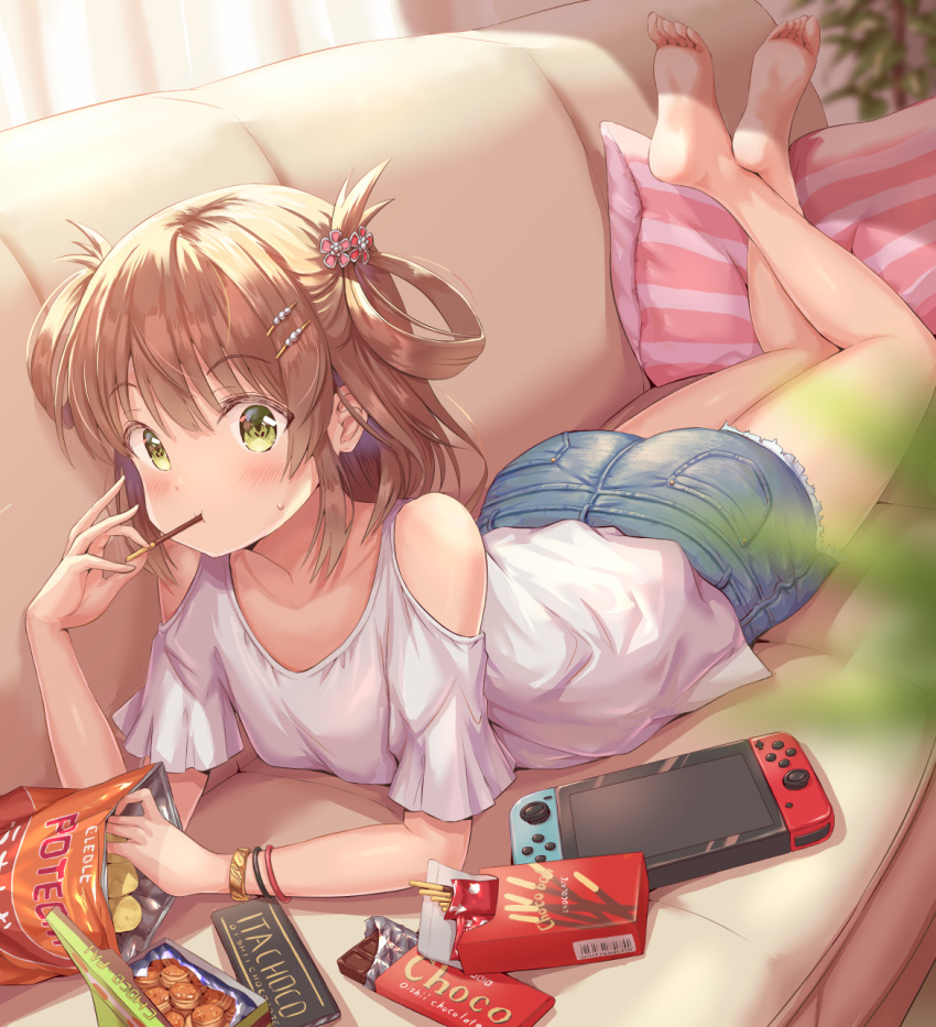 1girl bangs bare_shoulders barefoot blouse blush bracelet breasts brown_hair candy chips chocolate chocolate_bar collarbone couch crossed_legs denim denim_shorts eating eyebrows_visible_through_hair food green_eyes hair_ornament hair_rings hairpin handheld_game_console highres hirokazu_(analysis-depth) jewelry looking_at_viewer lying nintendo_switch no_shoes on_stomach pillow pocky potato_chips princess_connect! princess_connect!_re:dive rino_(princess_connect!) shorts small_breasts soles solo two_side_up white_blouse wristband