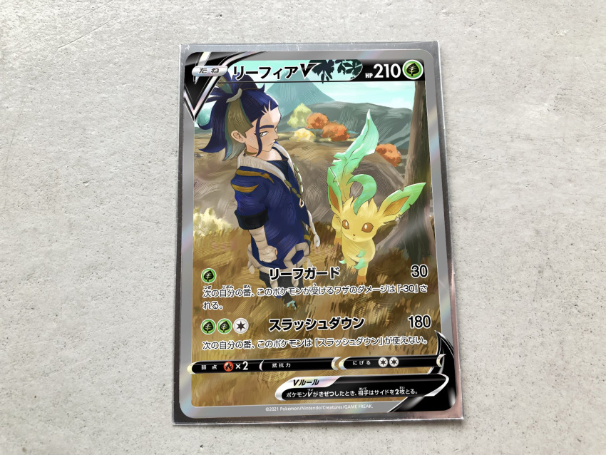 1boy adaman_(pokemon) arm_wrap blue_coat blue_hair card card_(medium) character_name character_print clenched_hand closed_mouth coat commentary_request day grass green_hair highres leafeon male_focus mountain multicolored_hair official_style pokemon pokemon_(game) pokemon_legends:_arceus pokemon_tcg pokeyugami ponytail standing tree