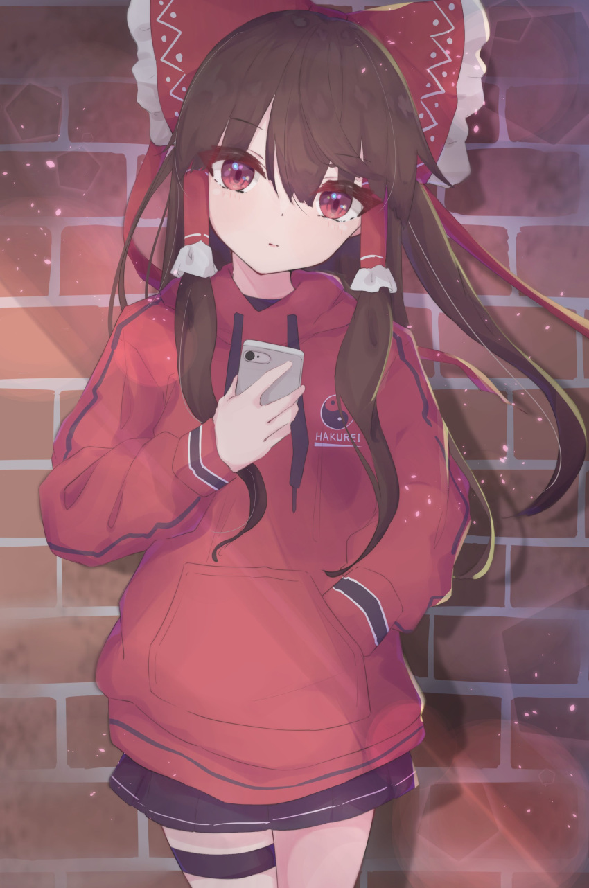 1girl absurdres alternate_costume black_skirt bow brick_wall brown_hair closed_mouth commentary contemporary hair_bow hair_tubes hakurei_reimu hand_in_pocket highres holding holding_phone hood hoodie long_hair long_sleeves looking_at_viewer matcha_yado phone red_bow red_eyes red_hoodie sidelocks skirt solo standing touhou yin_yang yin_yang_print