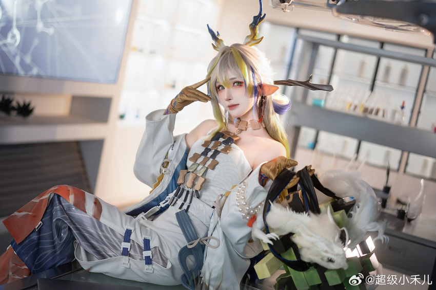 1girl arknights bare_shoulders blonde_hair blue_hair branch brown_gloves chaojixiaoheer closed_mouth collarbone commentary_request cosplay dragon_horns earrings eyeshadow gloves green_eyes grey_hair grey_pants highres holding horns indoors jacket jewelry laboratory long_hair long_sleeves looking_at_viewer makeup multicolored_hair necklace open_clothes open_jacket pants photo_(medium) pointy_ears red_eyeshadow shirt shu_(arknights) shu_(arknights)_(cosplay) sitting skirt smile solo streaked_hair stuffed_animal stuffed_dragon stuffed_toy weibo_username white_jacket white_shirt white_skirt