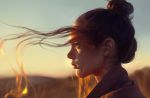  1girl artist_name astri_lohne_sjursen blurry brown_eyes brown_hair closed_mouth depth_of_field earrings freckles from_side hair_bun jewelry long_hair original outdoors portrait profile realistic sky solo sunset wind 