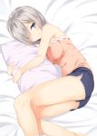  1girl bare_arms bare_shoulders bed bed_sheet blue_eyes blue_shorts breasts eyes_visible_through_hair hair_ornament hair_over_one_eye hairclip hamakaze_(kantai_collection) highres kantai_collection light_smile looking_at_viewer lying medium_breasts on_side one_eye_closed pillow pillow_hug pink_shirt shirt short_hair shorts silver_hair smile solo takehana_note tank_top thighs 