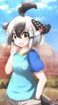  1girl ahoge arm_up bird_tail black_hair blue_shirt blue_sky blurry blurry_background clenched_hand clothes_writing clouds commentary_request day desert fang greater_roadrunner_(kemono_friends) green_eyes hand_in_pocket highres kemono_friends multicolored_hair open_mouth outdoors shirt short_sleeves shorts sky solo standing t-shirt talented_feather two-tone_hair upper_body upper_teeth white_hair 