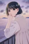  1girl bangs black_hair blurry blurry_background blush brown_dress closed_mouth depth_of_field dress eyebrows_behind_hair frilled_shirt frilled_sleeves frills from_side grey_eyes hand_to_own_mouth hand_up long_sleeves looking_at_viewer looking_to_the_side mattaku_mousuke original outdoors pleated_dress railing shirt sleeveless sleeveless_dress smile solo sunset thick_eyebrows twitter_username water white_shirt 