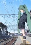  1girl bangs black_jacket black_legwear boots building commentary day from_side furuyama_itaru green_eyes green_hair hair_ornament hands_in_pocket hatsune_miku jacket kneehighs light_blush long_hair looking_to_the_side outdoors power_lines railroad_tracks solo standing tactile_paving train_station twintails very_long_hair vocaloid 