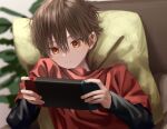  1boy aimai_sheena brown_hair brush_stroke closed_mouth faux_traditional_media from_above game_console hair_between_eyes holding indoors long_sleeves looking_at_object lying male_child male_focus nintendo_switch on_back original pillow red_eyes red_shirt shirt short_hair solo symbol-only_commentary 