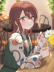  1girl blurry blurry_background blush brown_hair commentary_request cup disposable_cup dress flower green_dress highres holding holding_cup idolmaster idolmaster_shiny_colors indoors long_sleeves looking_at_viewer medium_hair osaki_tenka patterned_clothing pinafore_dress sitting sleeveless sleeveless_dress smile solo twitter_username white_flower yellow_flower yumeme_818 