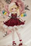  1girl adapted_costume belt blonde_hair blurry blurry_background brown_belt character_doll crystal depth_of_field doll fangs flandre_scarlet frilled_skirt frills full_body hat head_tilt highres medium_hair miyako_sasara mob_cap multicolored_wings one_side_up open_mouth petticoat photo_(medium) puffy_short_sleeves puffy_sleeves red_eyes red_skirt red_vest shirt short_sleeves skirt solo standing touhou vest white_headwear white_shirt wings wrist_cuffs 
